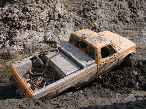 2000 Ford F-450 - Orange crew-cab Ford pickup buried to the door sills in deep mud at a pit, its flatbed carrying aluminum diamond-plate toolboxes and coils…