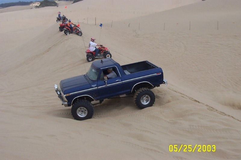 Blue lifted Ford Bronco driving on sand dunes with quad ATVs behind it