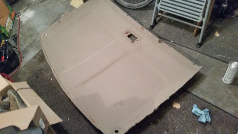 Car headliner removed and lying on a garage floor. It has a rectangular cutout for a light or speaker