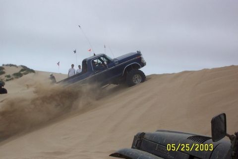 2000 Ford F-450 - A lifted dark blue late-1970s Ford pickup with a whip antenna and tall dune flag climbs a sand dune, spraying sand off its oversized tires…