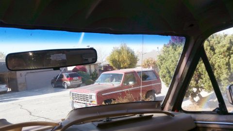 1985 Ford Bronco - View from inside a vehicle shows a red Ford Bronco parked on a residential street, with a garage and another SUV visible in the background.