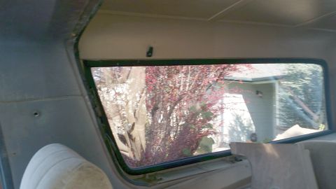 1985 Ford Bronco - Interior view of the rear passenger side window of a vehicle, looking out at a house and trees. The window trim and interior paneling are v…