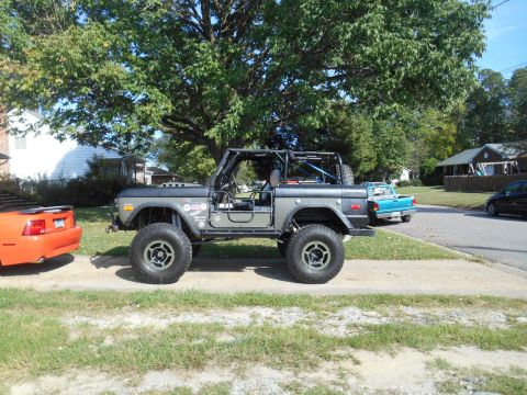 1972 Ford Bronco - A lifted, dark gray 1972 Ford Bronco is parked on a dirt and grass roadside next to an orange Ford Mustang. A turquoise pickup truck is par…