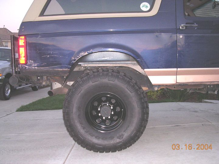 Rear quarter of a blue and tan lifted Ford Bronco on a black steel wheel and mud tire in a driveway
