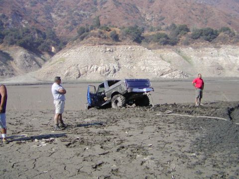 2000 Ford F-450 - A blue doorless lifted pickup with an open bed and oversized mud tires sits bogged down in a gray mud hole on a cracked dry lakebed, its bo…