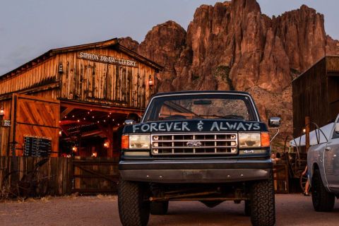 1988 Ford Bronco - A dark blue Ford F-Series truck is parked in front of a rustic wooden building with mountains in the background.