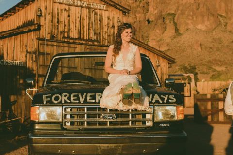 1988 Ford Bronco - A bride sits atop a black Ford truck, its hood painted with 'FOREVER WAYS', in front of a rustic wooden building.