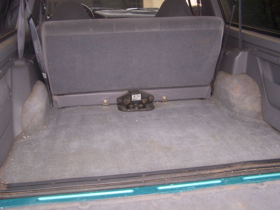 Interior cargo area of a vehicle with gray carpet and a folded-down rear seat. A black trailer hitch mechanism is visible