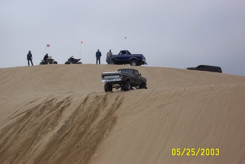Lifted trucks and ATVs on sand dunes under gray sky, blue Bronco at the crest