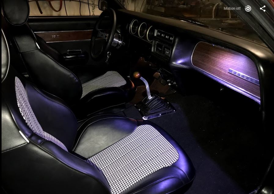 Interior of a classic car with black leather seats and houndstooth inserts, wood grain dash, and Hurst shifter.