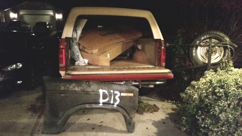 1985 Ford Bronco - A dark fender with white spray paint markings "80B 072" and "D13" rests on the ground in front of a red and tan SUV with its tailgate open,…
