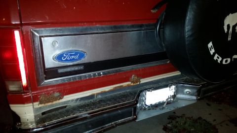 1985 Ford Bronco - The rear of a red 1985 Ford Bronco shows rust damage around the bumper and tailgate, with a spare tire cover visible.