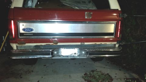 1985 Ford Bronco - The rear of a red and white Ford Bronco is shown with a chrome bumper and a silver tailgate panel featuring the Ford logo. The vehicle appe…