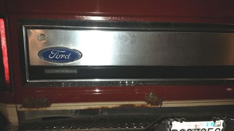 1985 Ford Bronco - The rear tailgate panel of a red Ford truck or SUV shows some rust damage along the bottom edge. A Ford oval emblem is visible on a brushed…