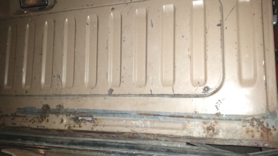 Close-up of a tan tailgate panel with vertical ribs and some rust near the bottom. Appears to be from an older Ford Bronco