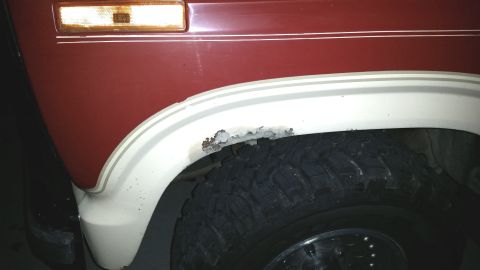 1985 Ford Bronco - Detail of a red SUV fender showing rust damage on the white fender flare, with a portion of the tire visible.