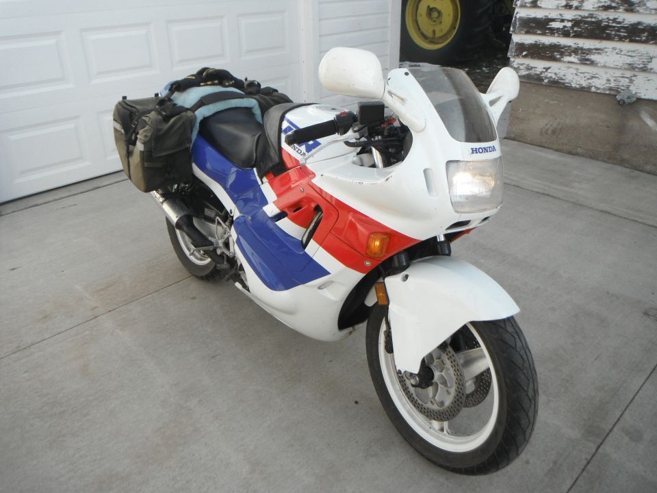 White Honda sport bike with red/blue graphics, saddlebags and sheepskin seat, parked on driveway by garage doors