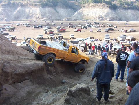 2000 Ford F-450 - A yellow Ford pickup lettered 'Tonka' and 'Pistons' crawling up a steep dirt obstacle at an off-road park, with a large crowd of spectators…