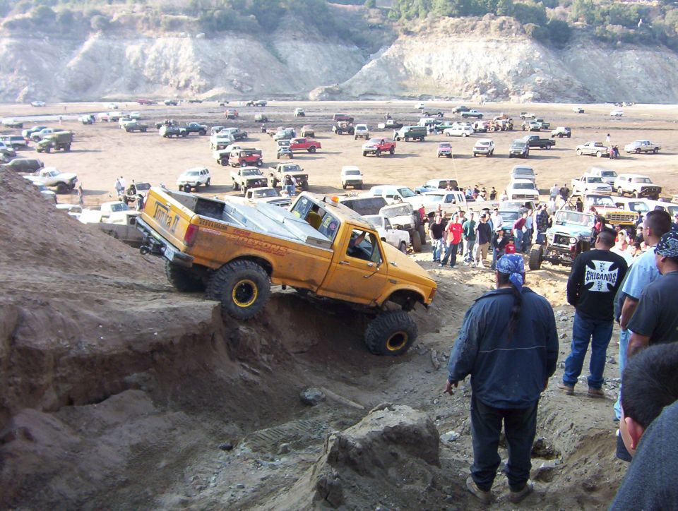 Yellow 'Tonka' Ford pickup climbing a steep dirt hill before a crowd at an off-road park