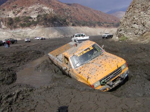 2000 Ford F-450 - A yellow/orange Ford Ranger-style pickup with a 'Tonka II' windshield banner sits buried to the axles in deep gray mud at an off-road park,…