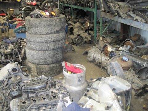 1989 Ford Bronco - Various car parts, including engines and tires, are piled up in a disorganized auto parts storage facility.