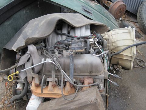 1989 Ford Bronco - A Ford V8 engine and transmission assembly, partially covered, sits in a junkyard setting.
