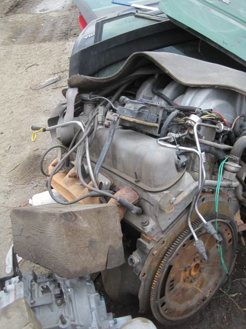 1989 Ford Bronco - Engine assembly with V8 intake manifold and exhaust manifold, supported by a wooden block in a junkyard setting.