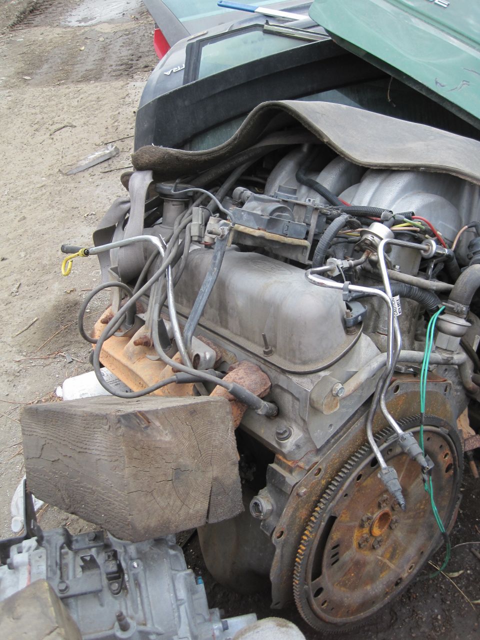 Engine with V8 intake manifold and exhaust manifold. A wooden block supports the engine