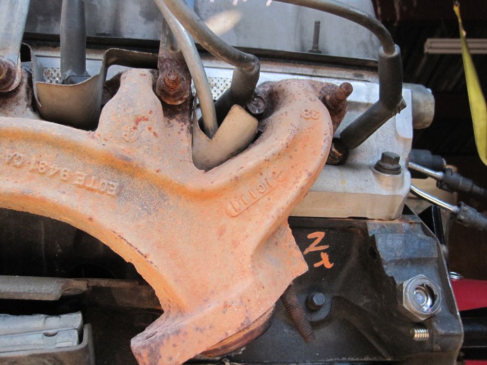 Close-up of a rusty exhaust manifold with '88' cast into it and 'E0TE 9483 CA' stamped on the pipe