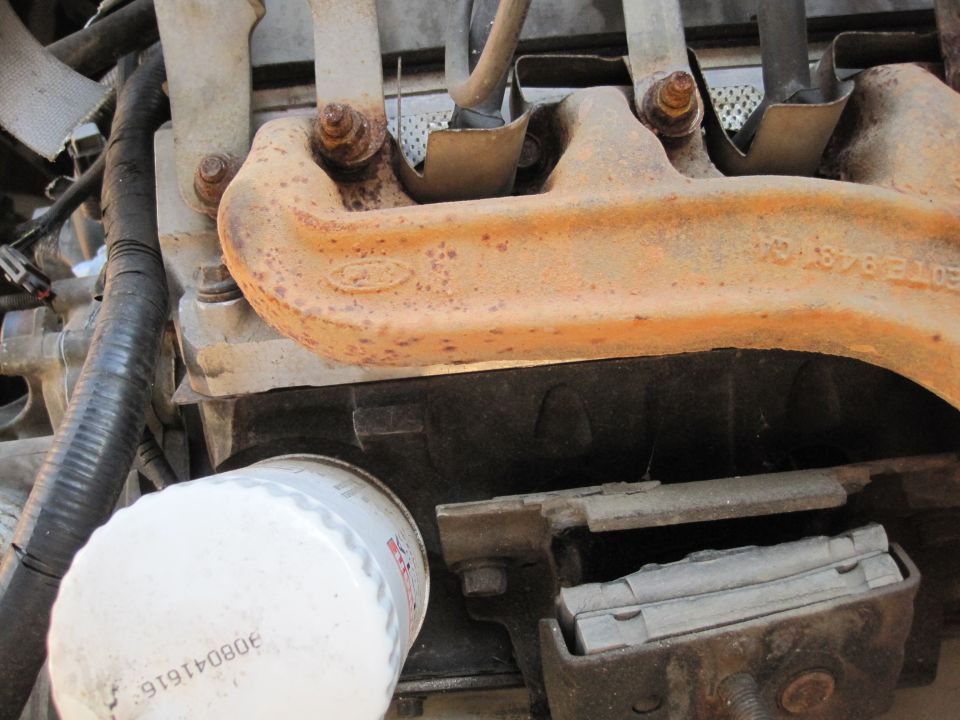 Close-up of a rusty Ford exhaust manifold with a Ford logo and part numbers visible. An oil filter is also in the shot