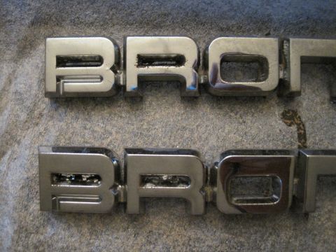 1989 Ford Bronco - Two chrome Bronco emblems are shown on a gray, textured surface. The text is clearly visible on both.