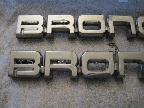 1989 Ford Bronco - Two chrome Bronco emblems are shown close-up on a gray, textured background.