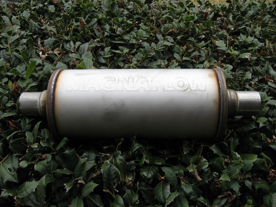 Magnaflow exhaust muffler with inlet and outlet pipes, resting on green foliage