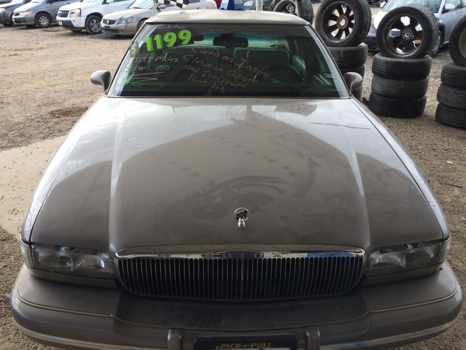 Gray sedan with a chrome grille and rectangular headlights, parked in a lot with other vehicles and stacked tires