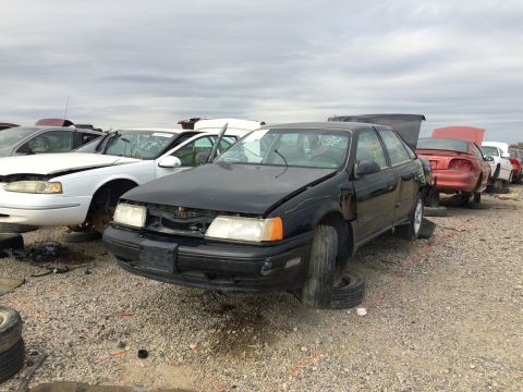 1985 Jaguar XJ6 - A black sedan is parked in a junkyard with its hood open and flat tires.