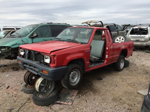 1985 Jaguar XJ6 - A red pickup truck sits in a junkyard, missing its grille and doors, with other vehicles visible in the background.