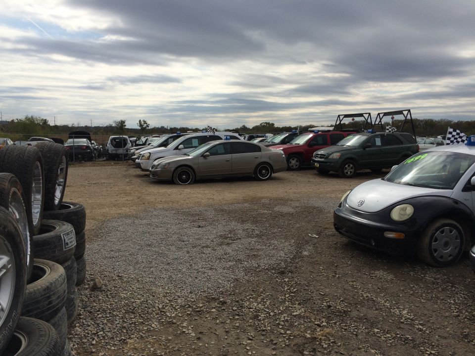 A car lot with many vehicles, stacks of tires, and a Volkswagen Beetle in the foreground.