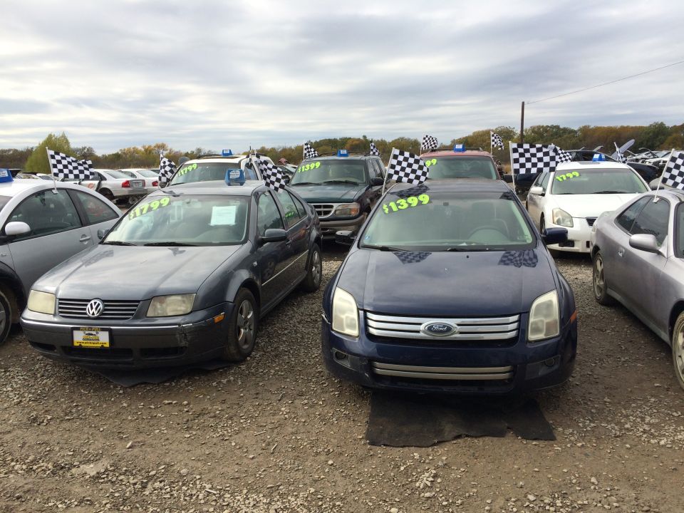 Dark blue Ford Fusion and gray Volkswagen sedan at a car lot with checkered flags and price tags.