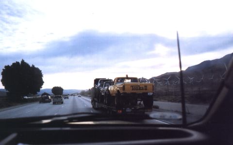 2000 Ford F-450 - Windshield-view shot down a freeway of a flatbed trailer hauling a yellow lifted Ford pickup with 'Tonka II' lettered on the tailgate, a se…