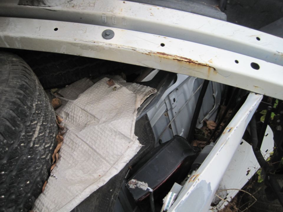 Close-up of damaged white vehicle undercarriage with tire, crumpled material, and rust.