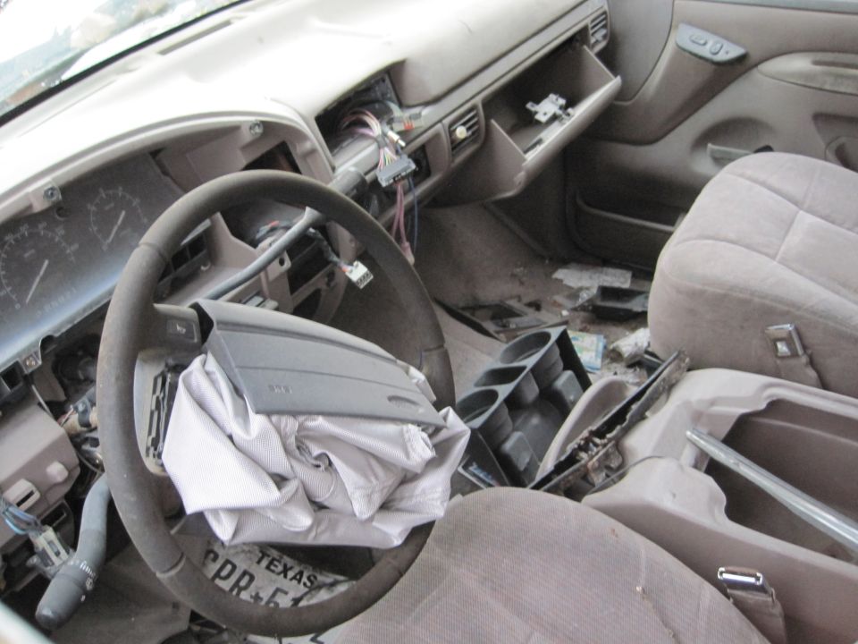 Interior of a damaged vehicle with deployed airbag, cracked dashboard, and Texas license plate on floor.