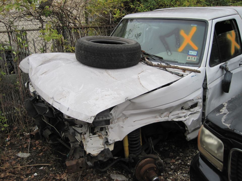 Damaged white pickup truck with spare tire on hood and Xs on windshield.