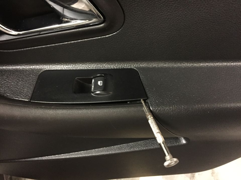 Car interior door panel with window switch trim being pried open by a screwdriver.
