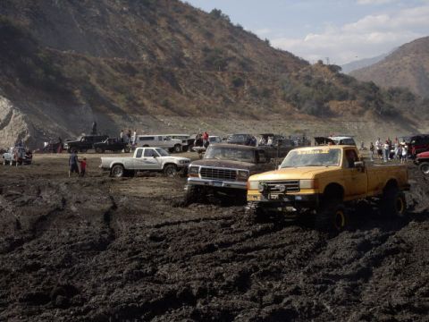 2000 Ford F-450 - Three pickups lined up at the edge of a churned mud pit in a canyon riding area: a lifted yellow 1990s Ford with windshield lettering and t…