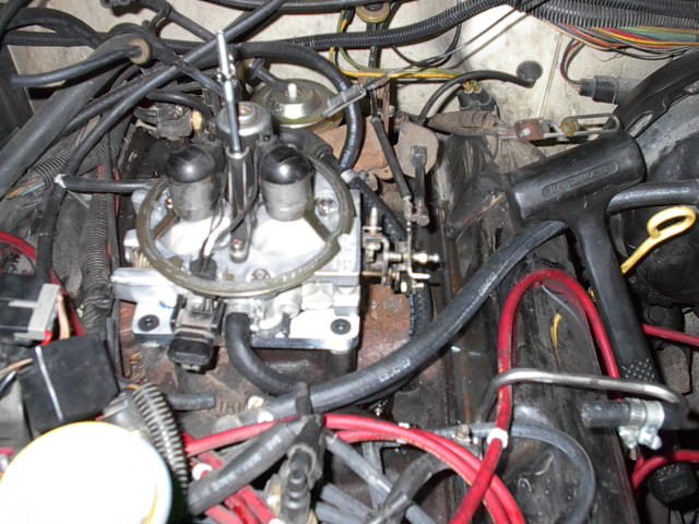 Engine bay with a carburetor, hoses, and wiring. A black rubber mallet rests on a black hose