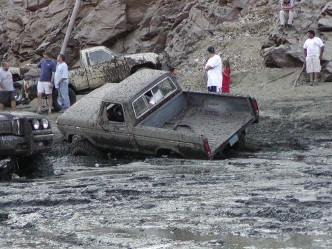 2000 Ford F-450 - A 1970s-era short-bed pickup, completely coated in gray mud with its doors removed, sits axle-deep in a mud pit at the edge of a rocky bank…