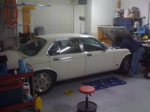 1987 Jaguar XJ6 - A person works on the engine of a cream-colored 1987 Jaguar XJ6 sedan parked in a garage.