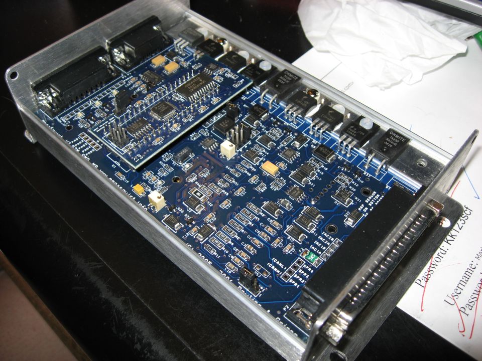 Circuit board with multiple integrated circuits, resistors, and capacitors in a metal casing. Text is visible on the board and on paper underneath