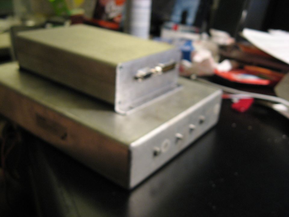 Two metal electronic enclosures stacked on a dark surface. The top enclosure has a connector on its side