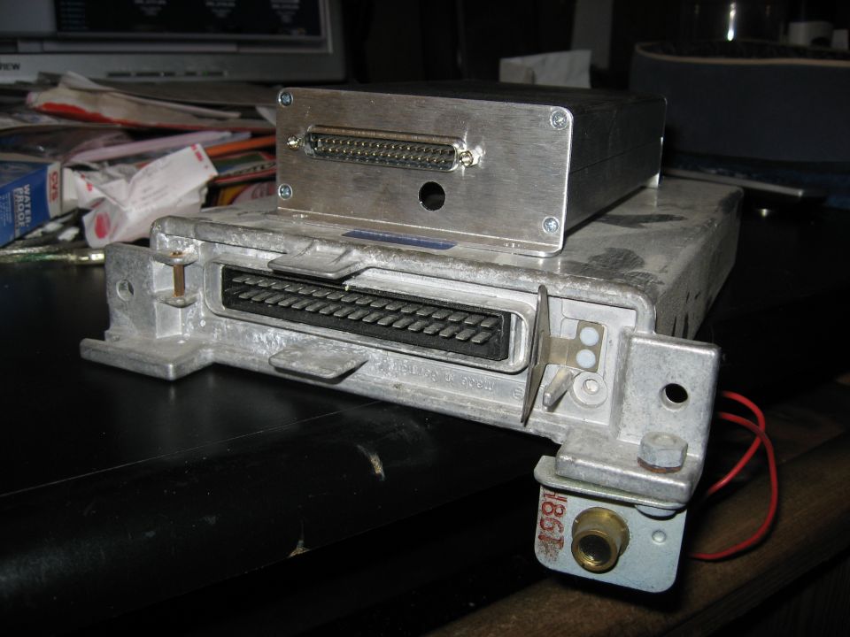 Two engine control units stacked, one on top of the other. The bottom unit has a large black connector. The top unit is a silver box with a smaller connector